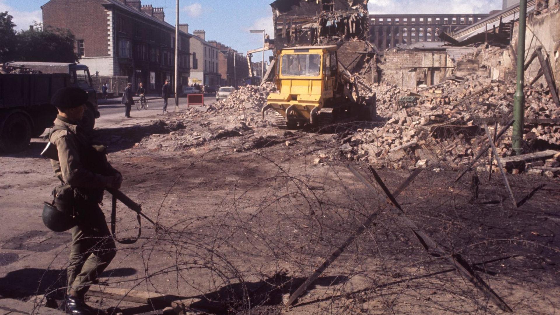 A still from 1969 of a soldier at a barbed wire barrier as there is a digger clearing up rubble on the Falls Road in Belfast.