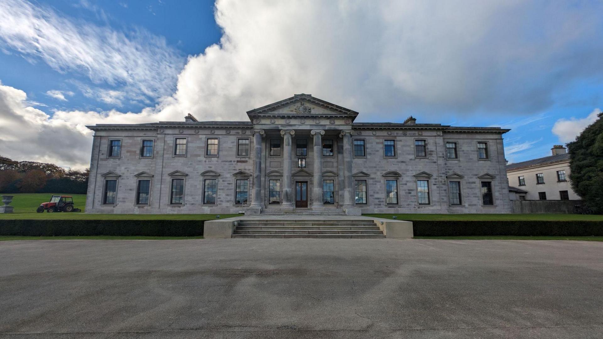  Ballyfin Demesne hotel - a grey stone building, with four pillars at the front of it, sits under a cloudy sky in Laois, Ireland in 2023. There is a large grassy area in front and behind it