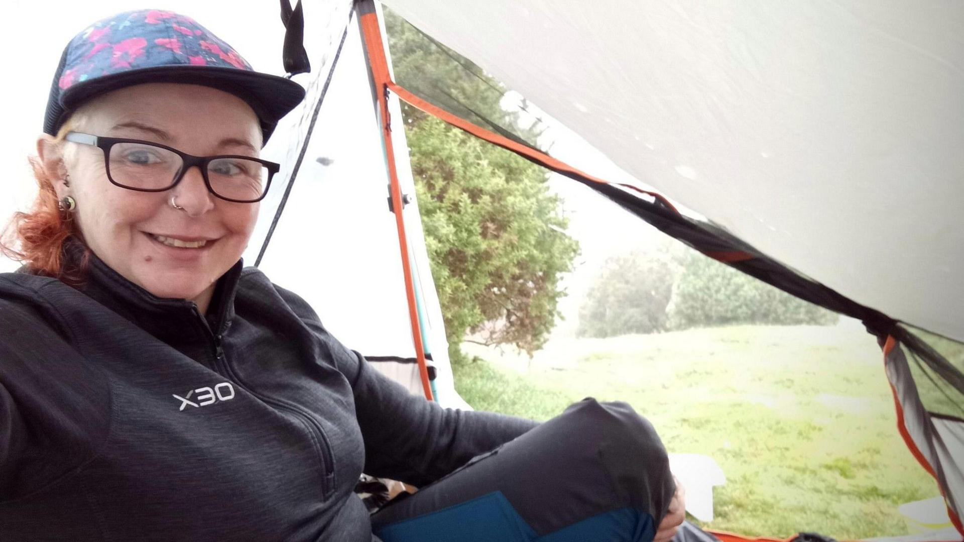Becky Harrison sitting inside a tent while wild camping. Ms Harrison is smiling at the camera. The tent door is open and is showing a field and trees.