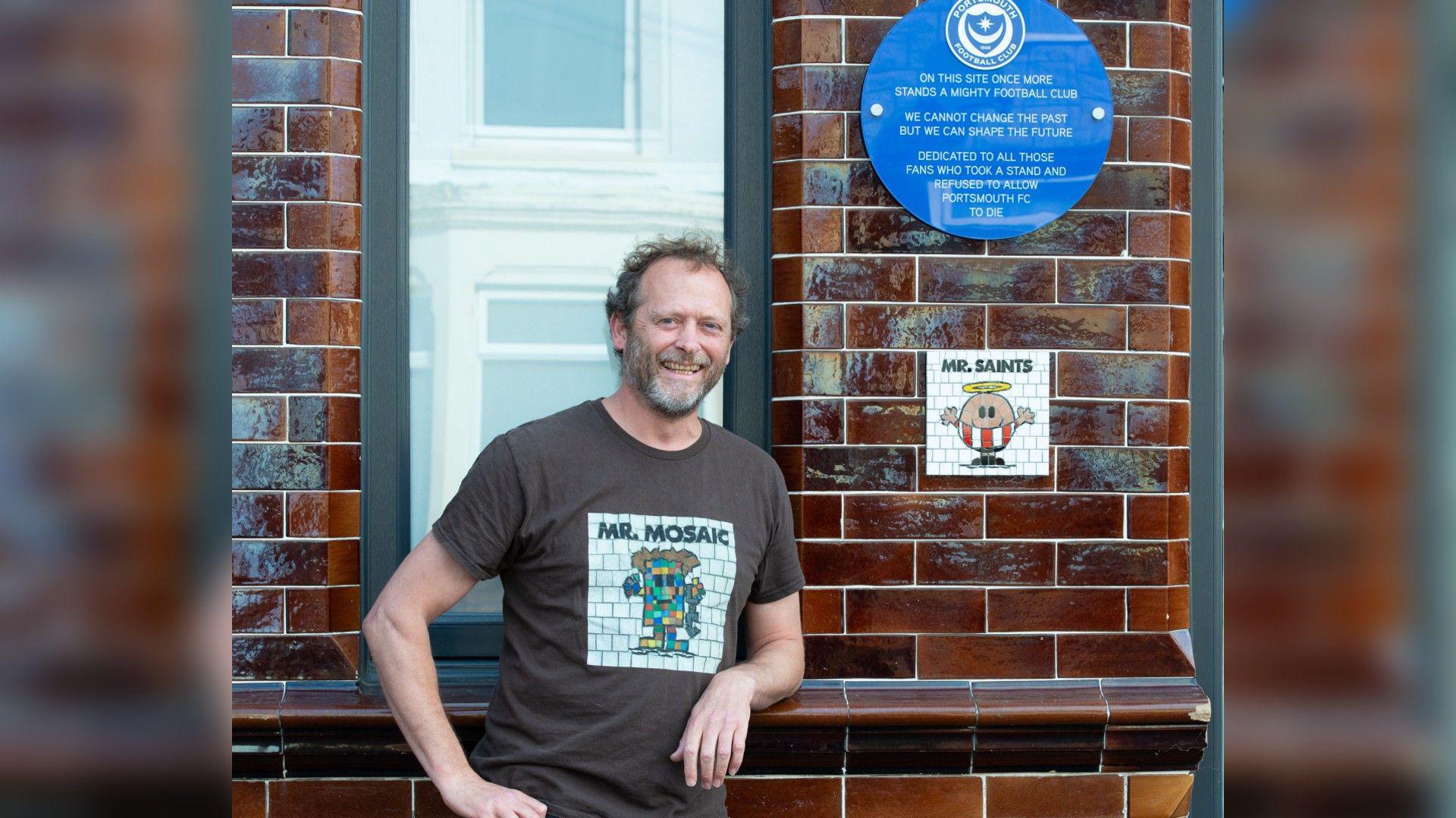 Will Rosie wearing a t-shirt which says "Mr Mosaic" he is leaning on the wall where there is a blue plaque which shows the Portsmouth Football Club crest above text which reads "on this site once more stands a mighty football club. We cannot change the past but we can shape the future. Dedicated to all those fans who took a stand and refused to allow Portsmouth FC to die" underneath the plaque is a white mosaic with a mr man character dressed in a southampton fc kit, it says "mr saints" at the top