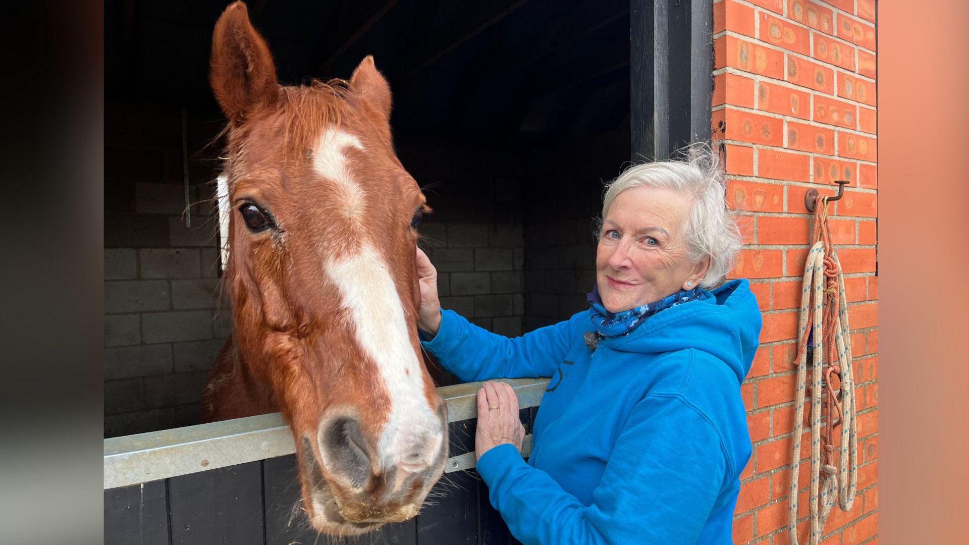 A woman with short white hair smiles gently as she looks at the camera. She wears a blue hooded top and pats a brown horse, who is hanging over its stable door.