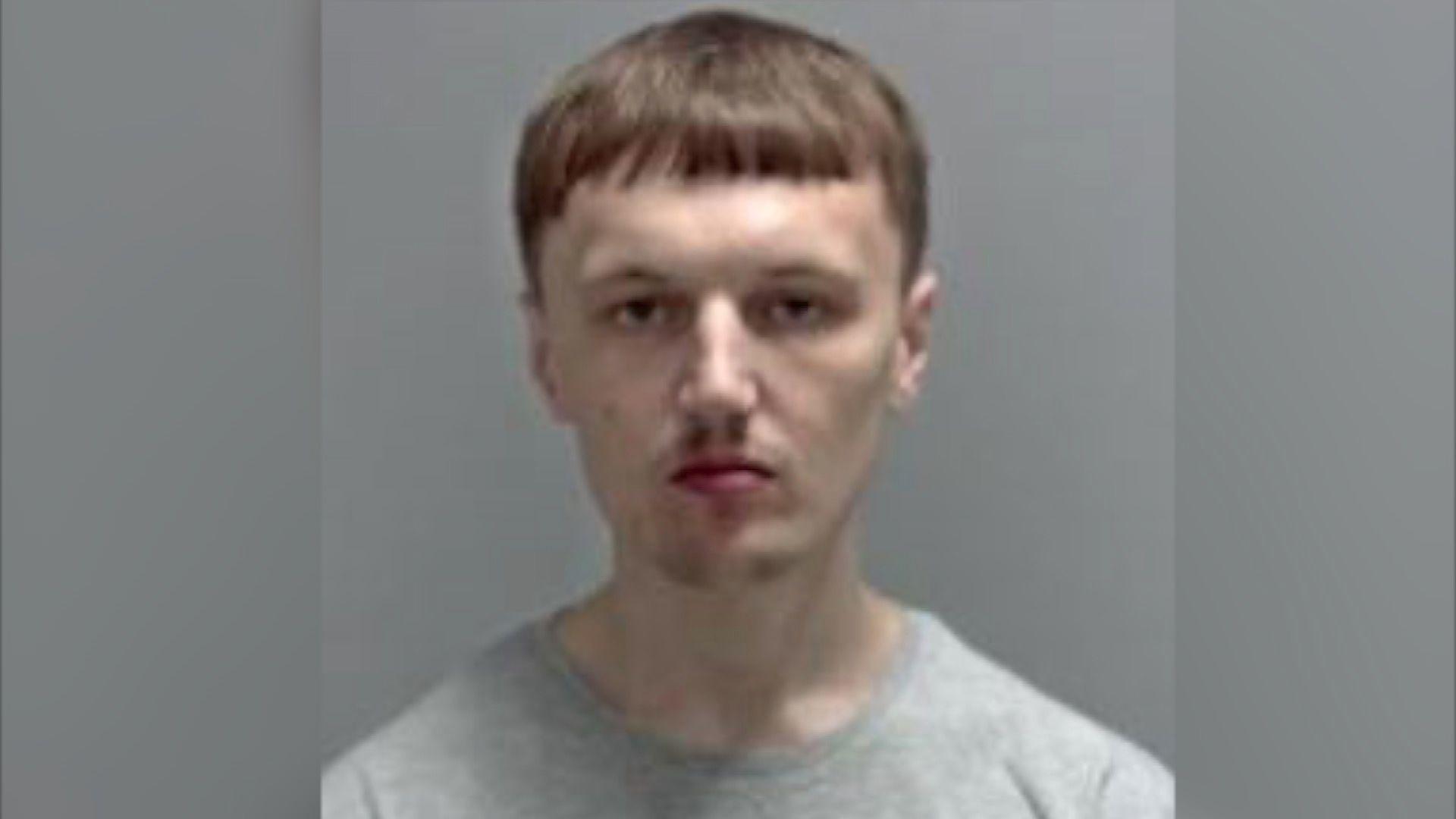 A custody image of Sam Morrison, who is wearing a grey top, and has a short brown haired fringe. He has a slight moustache.