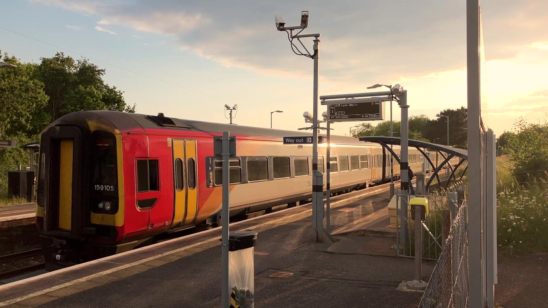 A South Western Railway train travelling through a station past an empty platform on a sunny day.