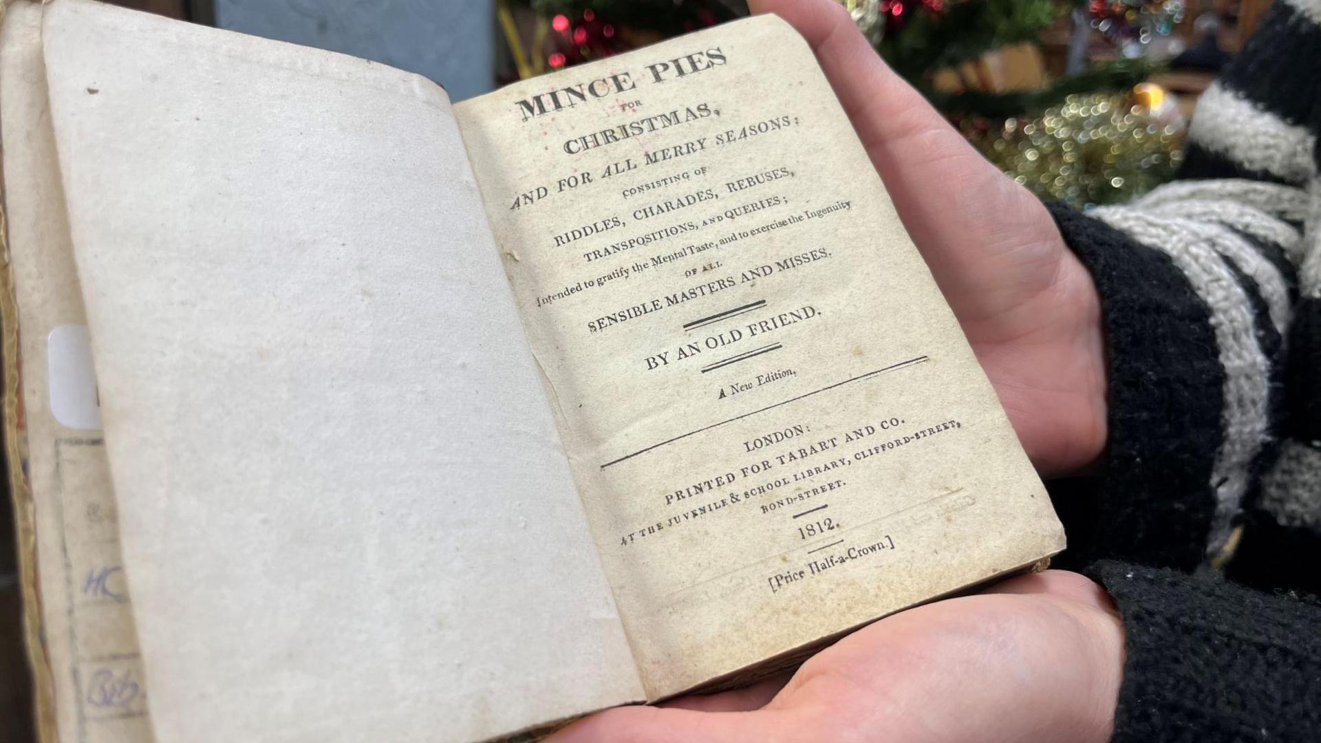 A close-up picture of an old book which reads Mince Pies at Christmas. The book is being held in someone's hands. 