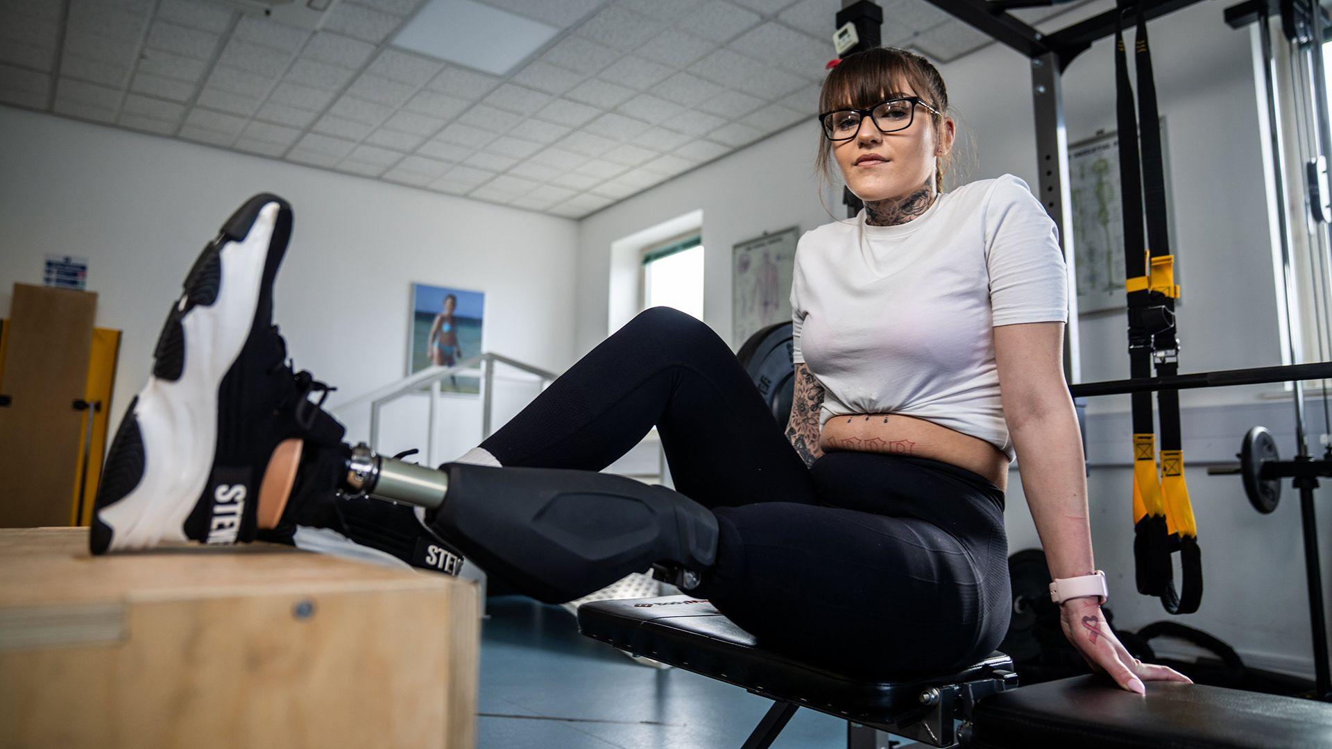 A woman in a white t-shirt and black trousers, with her feet up on a wooden box. One of her legs is a prosthetic leg with a trainer on the foot.