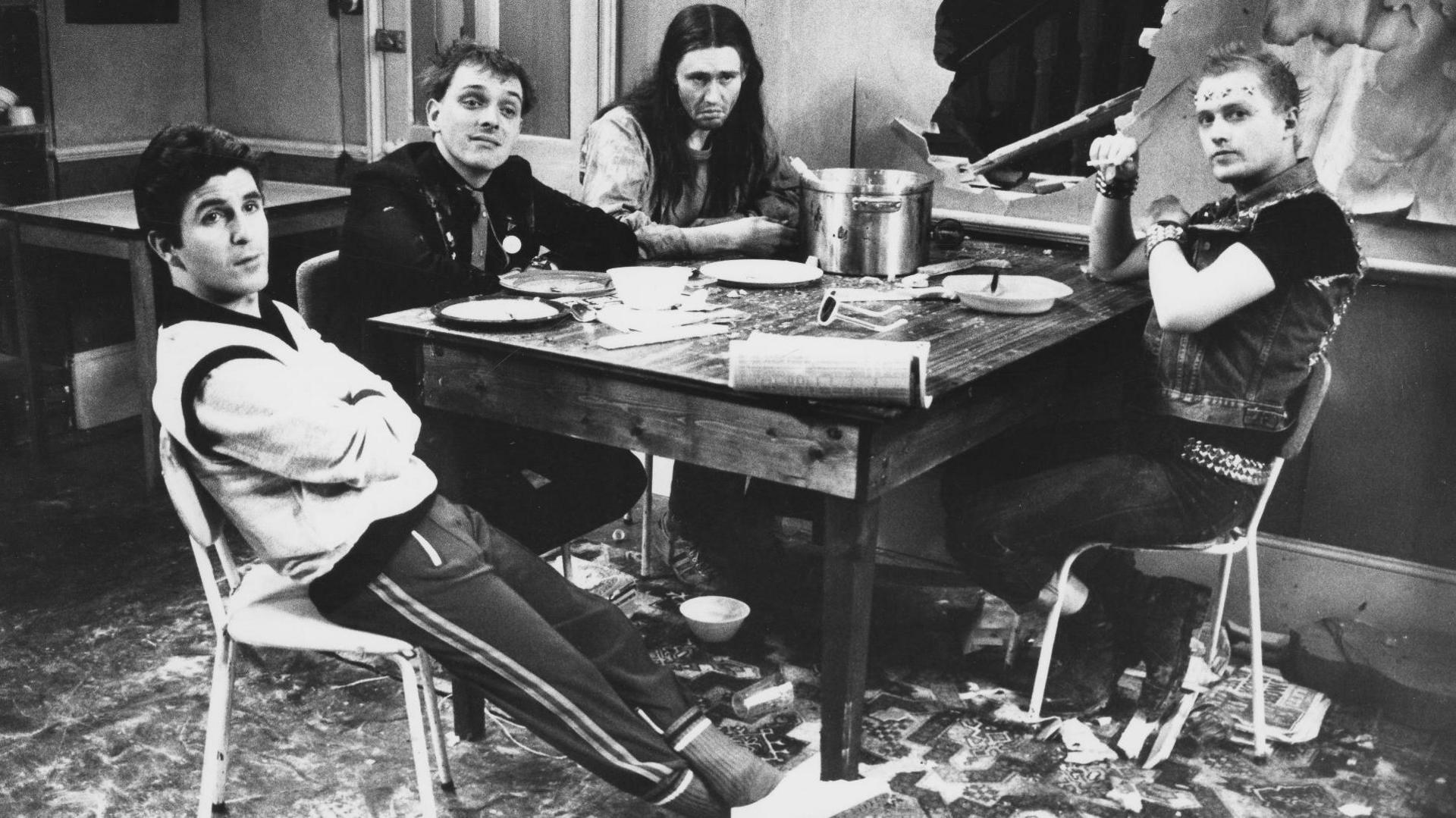 A black and white photo of four white males - Christopher Ryan, Rik Mayall, Nigel Planner and Adrian Edmondson - sitting around a table in a scene from the television sitcom 'The Young Ones'