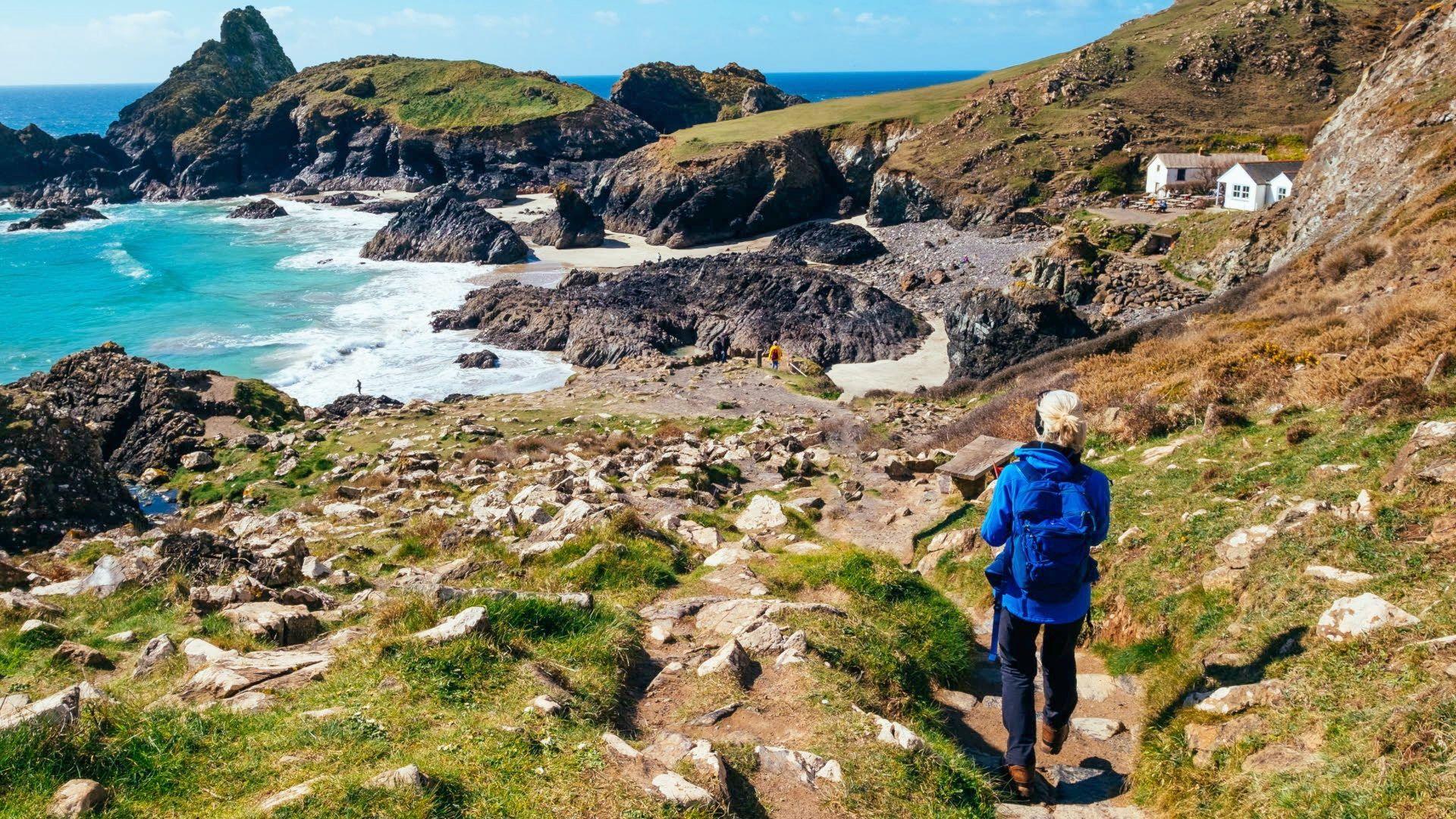 Woman walking on a footpath towards the sea. The path winds down dramatic, rocky cliffs. 