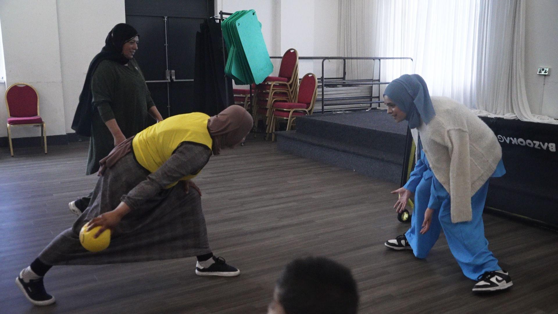 Three women pictured in traditional Muslim dress. One is trying to throw a ball past another to score a goal. 