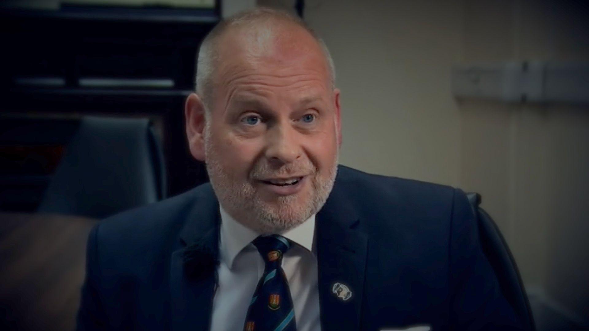 Jonathan Nunn sitting close to the camera indoors, dressed in a navy blue suit and striped tie. He appears with receding white hair and stubble.