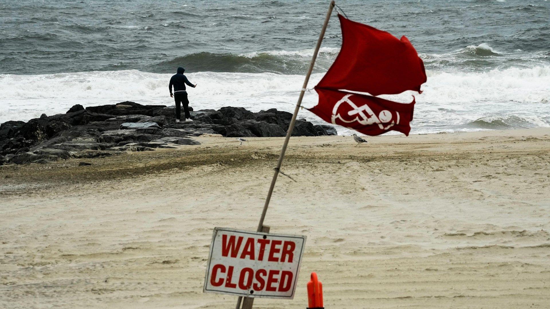 Man on an otherwise empty beach that shows large waves and a red flag and a sign in the foreground that says 'water closed'.
