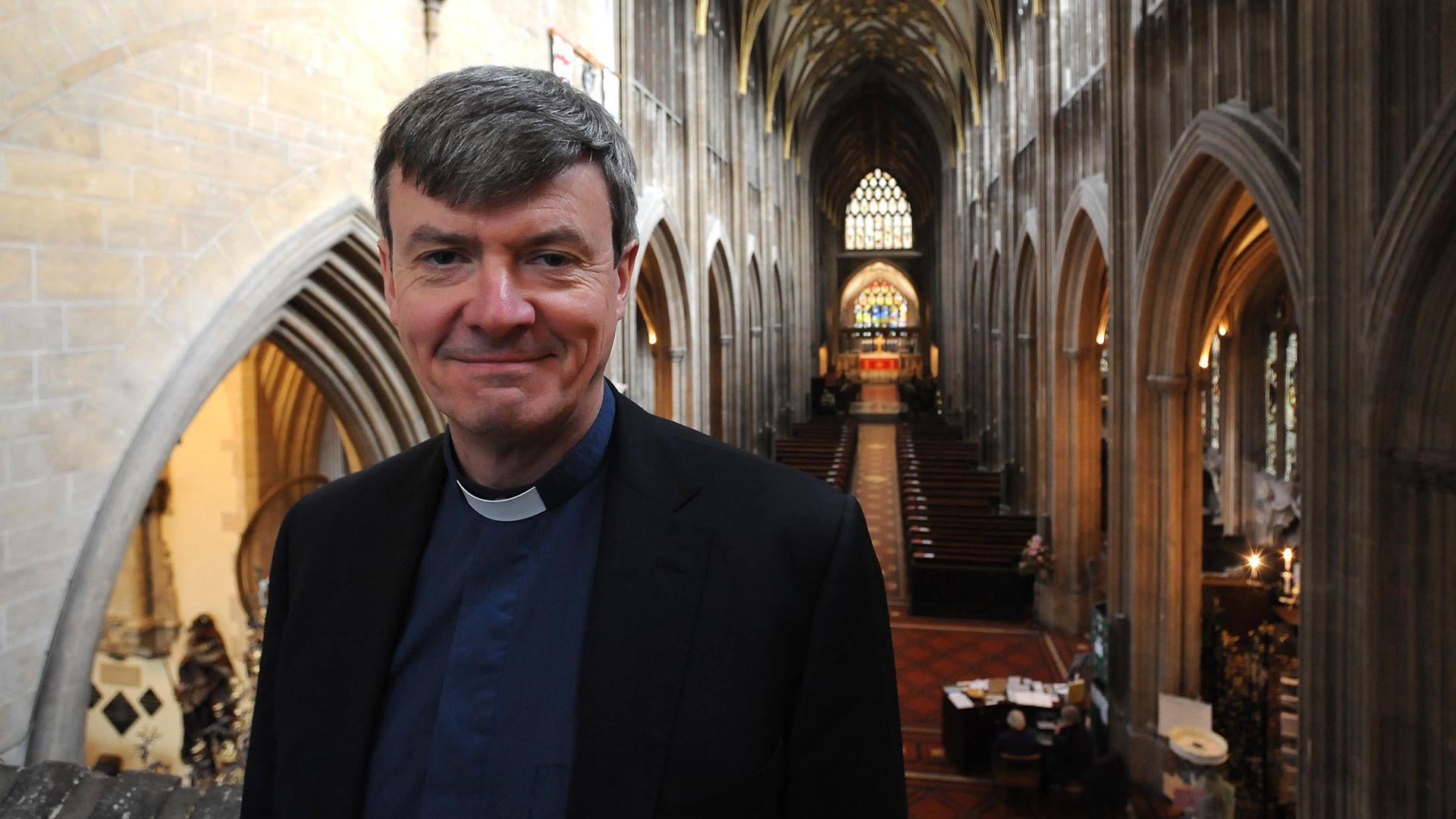 Dan Tyndall wearing a black blazer, a navy blue shirt and his vicar dog collar. He is smiling at the camera while standing on a balcony, overlooking the main hall of St Mary Redcliffe Church. There are archways on either side of the aisle and a large stained glass window at the far end.