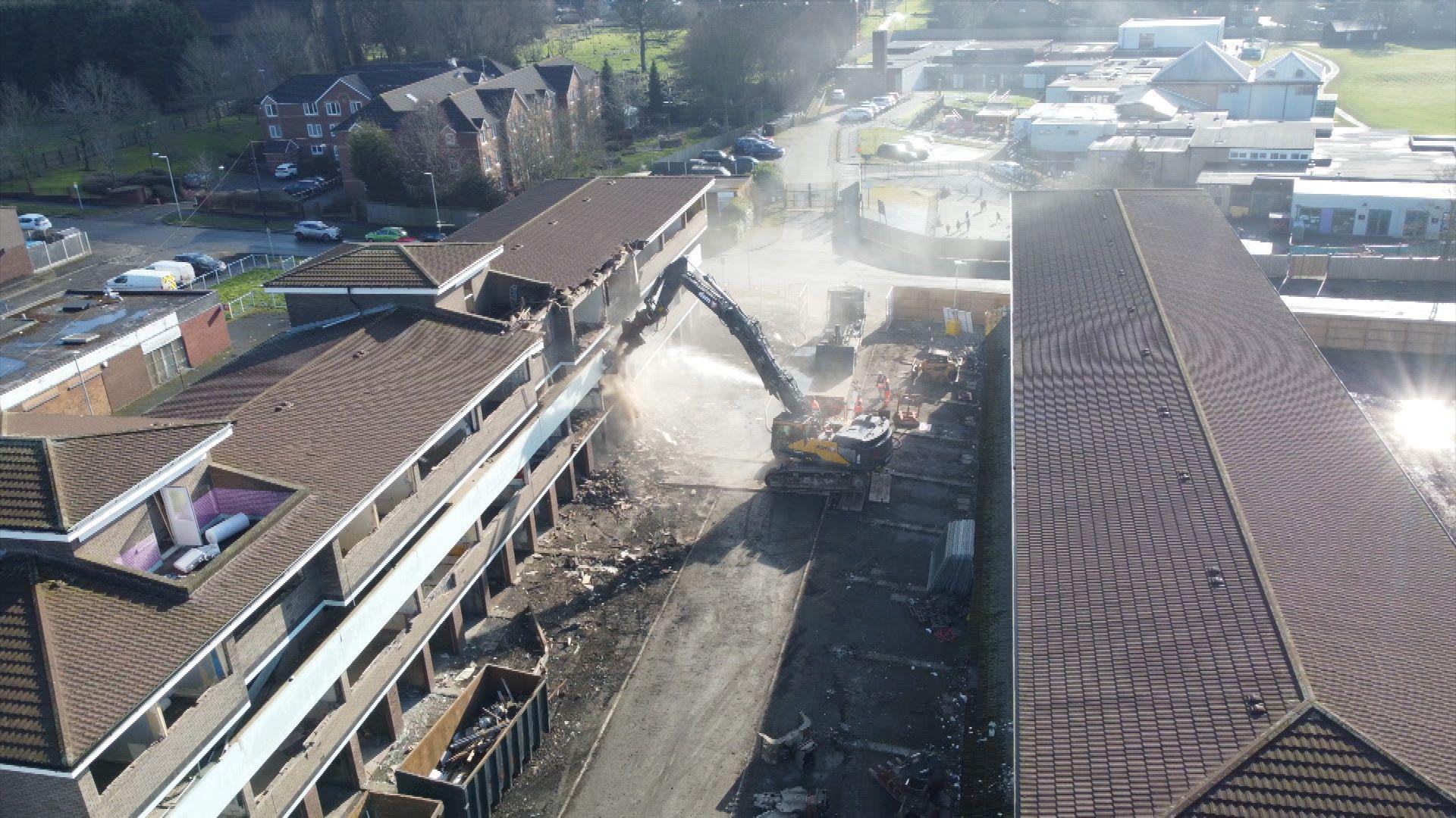 An aerial view of two blocks of the maisonettes. A digger begins demolishing one of them.