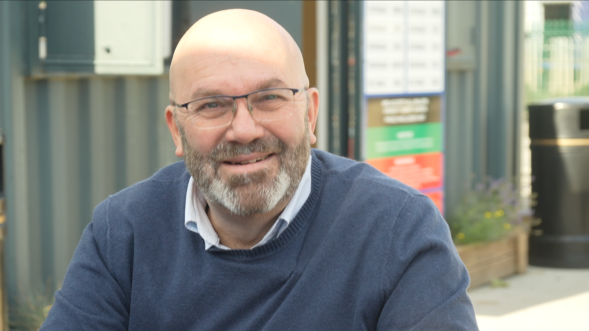 Man with glasses wears a shirt and blue jumper. He is smiling at the camera.