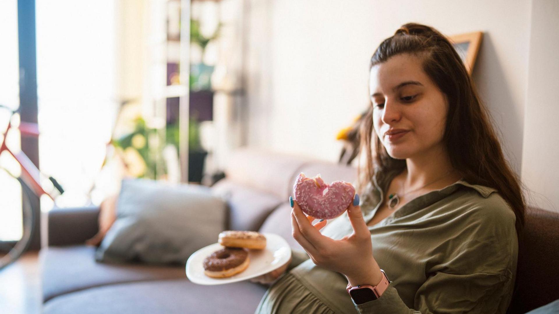 Pregnant woman sits on a sofa holding a plate of doughnuts with pink and chocolate-toppings. The pink doughnut is half-eaten.