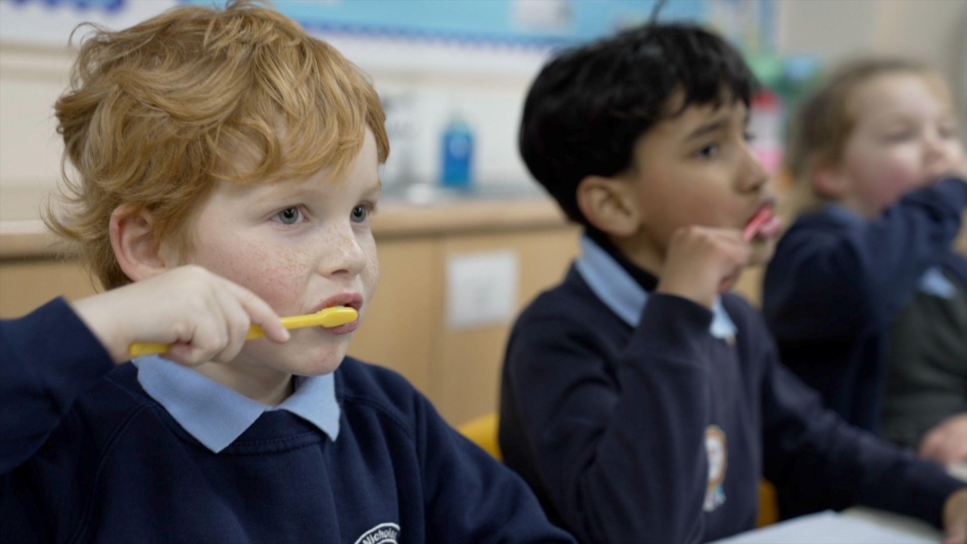 Three children are seen sat at their classroom desks brushing their teeth with multicoloured toothbrushes. They are wearing their school uniforms - a navy jumper with the collars of a light blue polo sticking out over the top