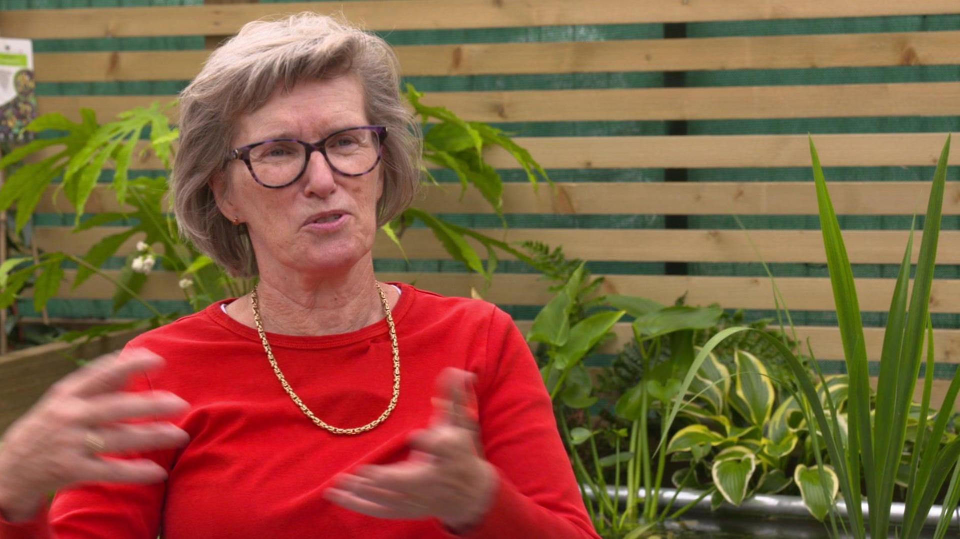 Angela Hayler, wearing a red top and a gold necklace, speaks to the camera