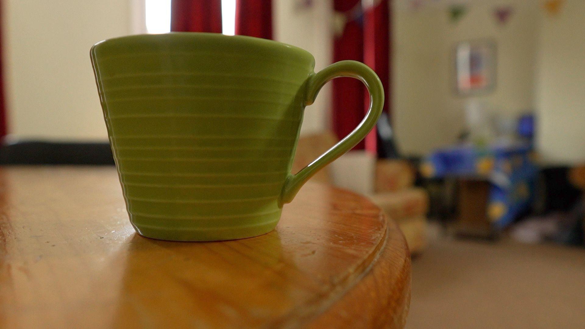 A green mug sitting on a wooden table. Windows, red curtains and a sofa can just be made out in the background.