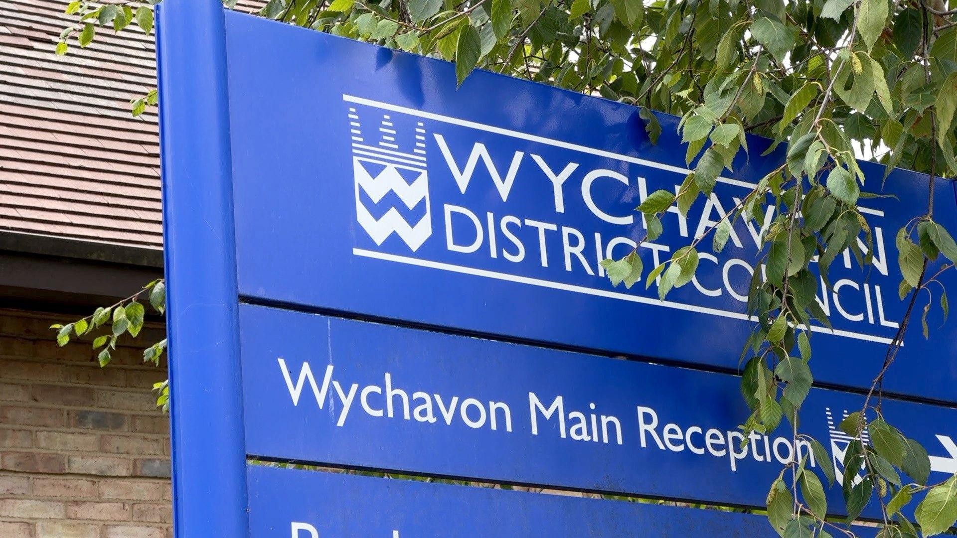 A blue sign outside a building, partly covered by leaves. The sign reads Wychavon District Council. Another sign with the words Wychavon Main Reception is directly underneath.