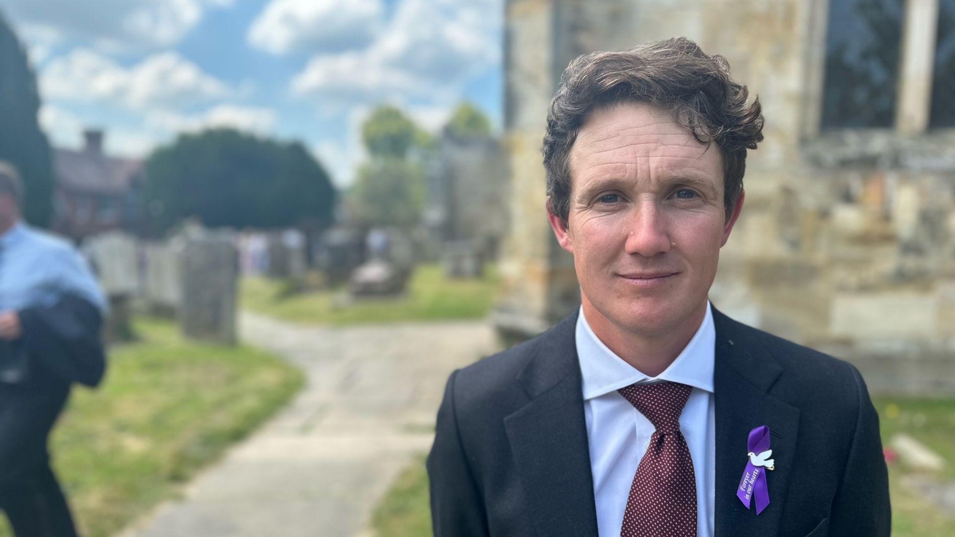 A man wearing a blue shirt, red tie and dark blue jacket stood in a church yard. The man has brown hair, he is looking at the camera. On his suit jacket is a purple and white ribbon.