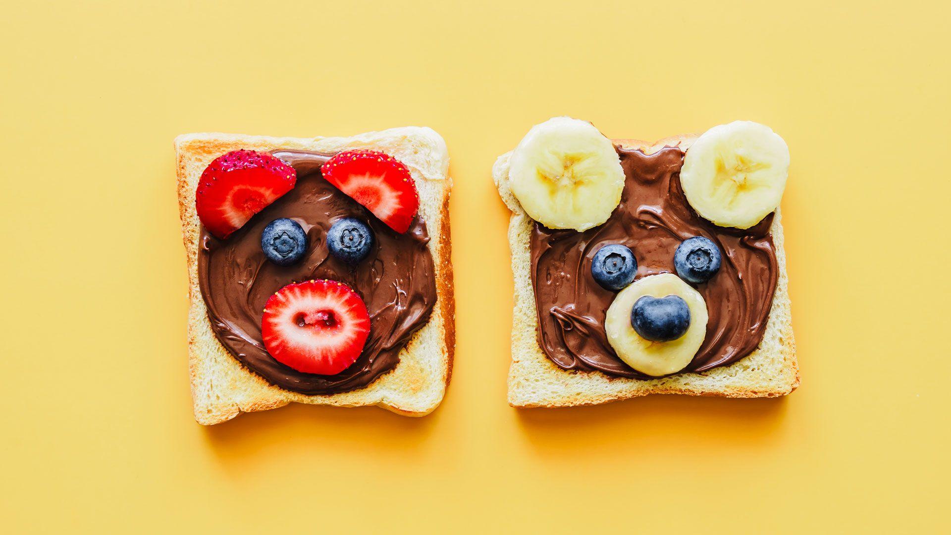 Two slices of toast with chocolate spread and fruit which is shaped to look like bears. The one on the left has strawberries for ears and mouth and blueberries for eyes and the one on the right has bananas for ears and mouth and blueberries for eyes and nose. On a yellow background