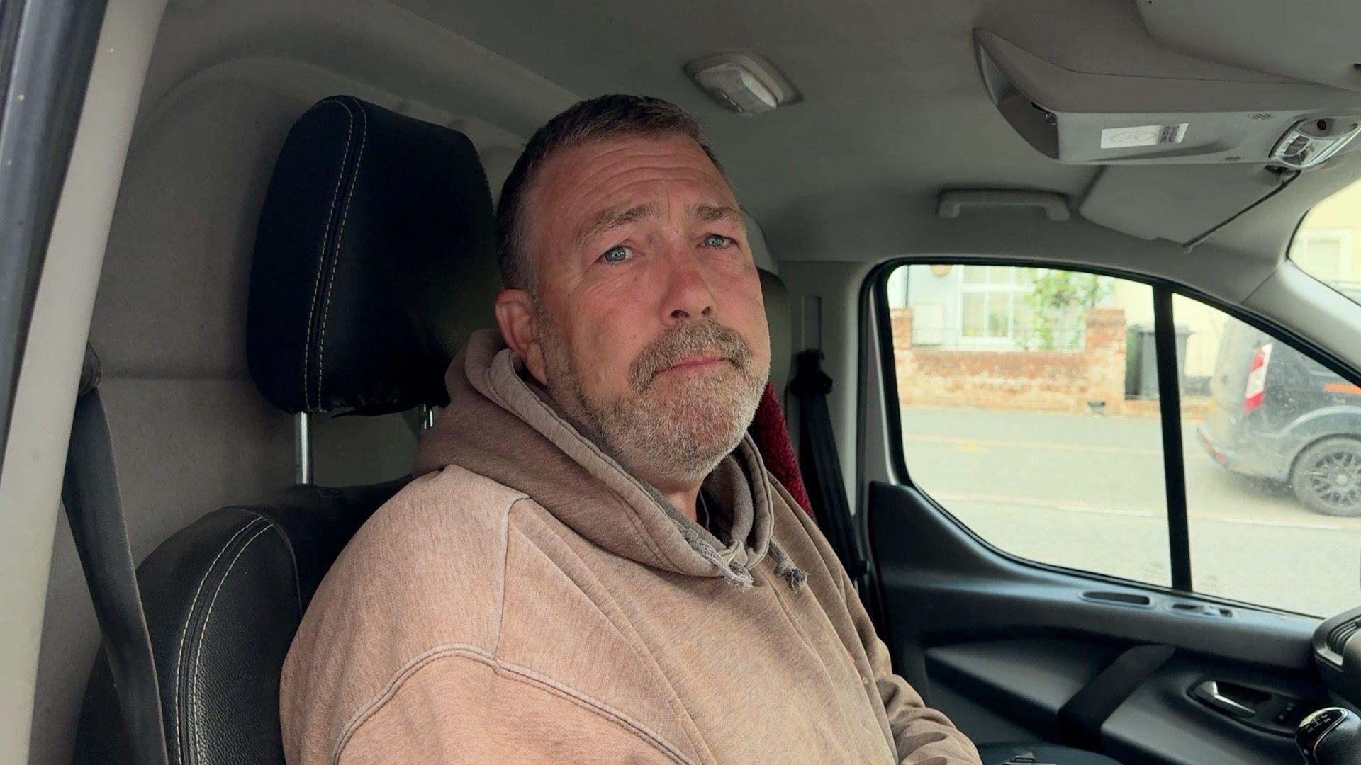 A man with short grey hair and beard , wearing a light brown hoodie, sitting in van.
