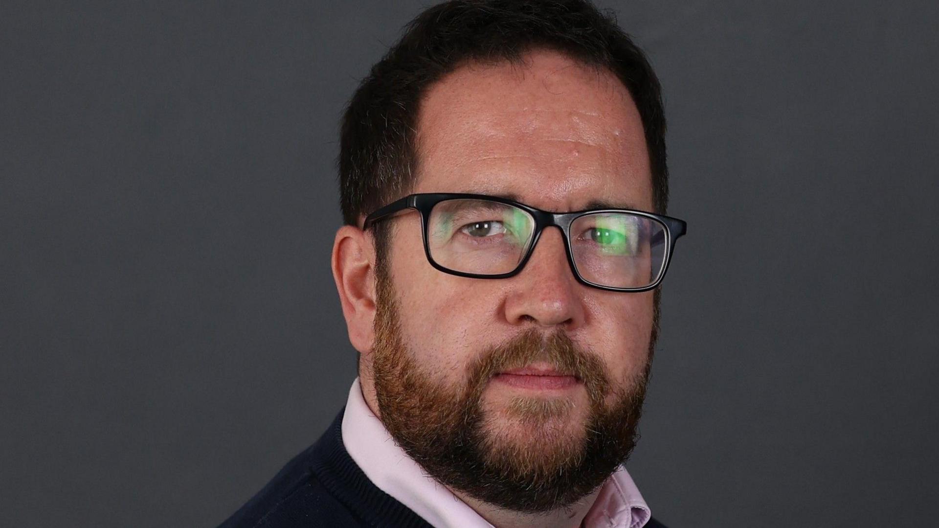 A dark-haired man with glasses, wearing a black top and pink shirt, looks at the camera