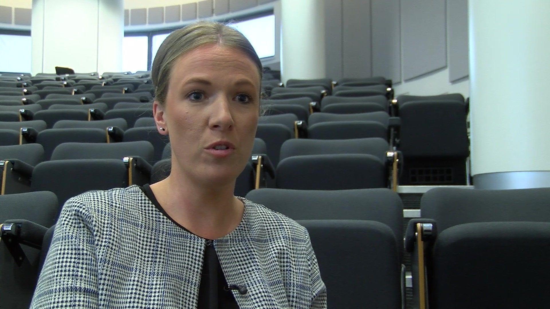 A young blonde woman in a checked blazer addressing the camera with an empty lecture theatre behind her