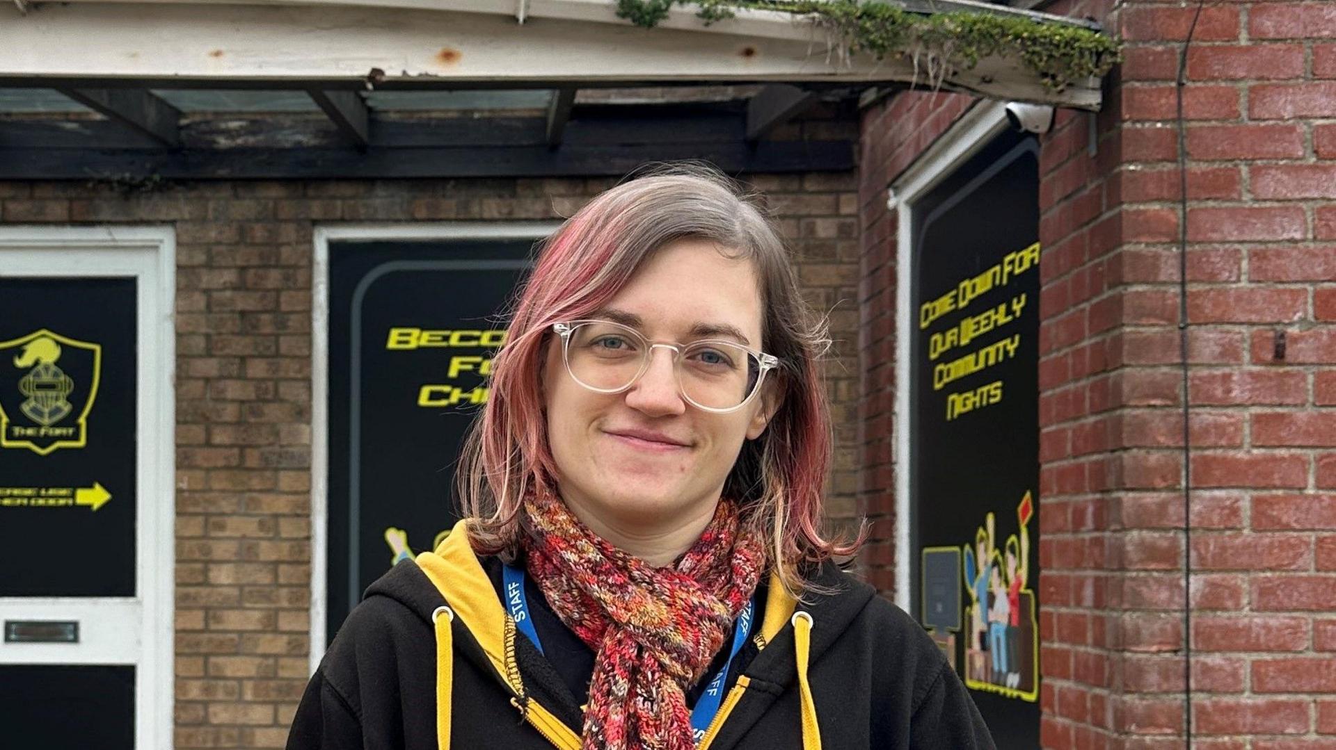 Giselle Czajka stands outside The Fort building. She has pink dyed hair with dark roots. She is wearing clear framed glasses with a predominantly red scarf tied around her neck. She is wearing a black hoodie with yellow strings and a yellow hood and a Staff ID badge under her scarf. 