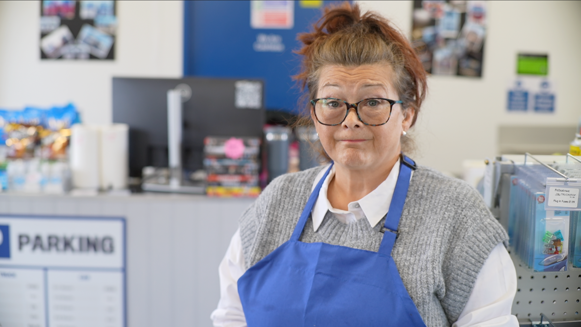 Woman with glasses and a blue apron over a grey sleeveless jumper and white shirt. She is standing inside the truck stop.