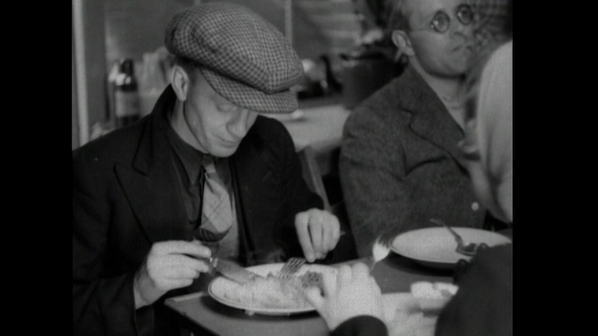 A black and white news clip of a man eating a meal at a table with a knife and fork. He is wearing a black overcoat, a dark shirt and a lighter coloured tie. He is also wearing a tweed-style checked cap and looking down at his food. A man to his left has an empty plate in front of him and is looking off into the distance.