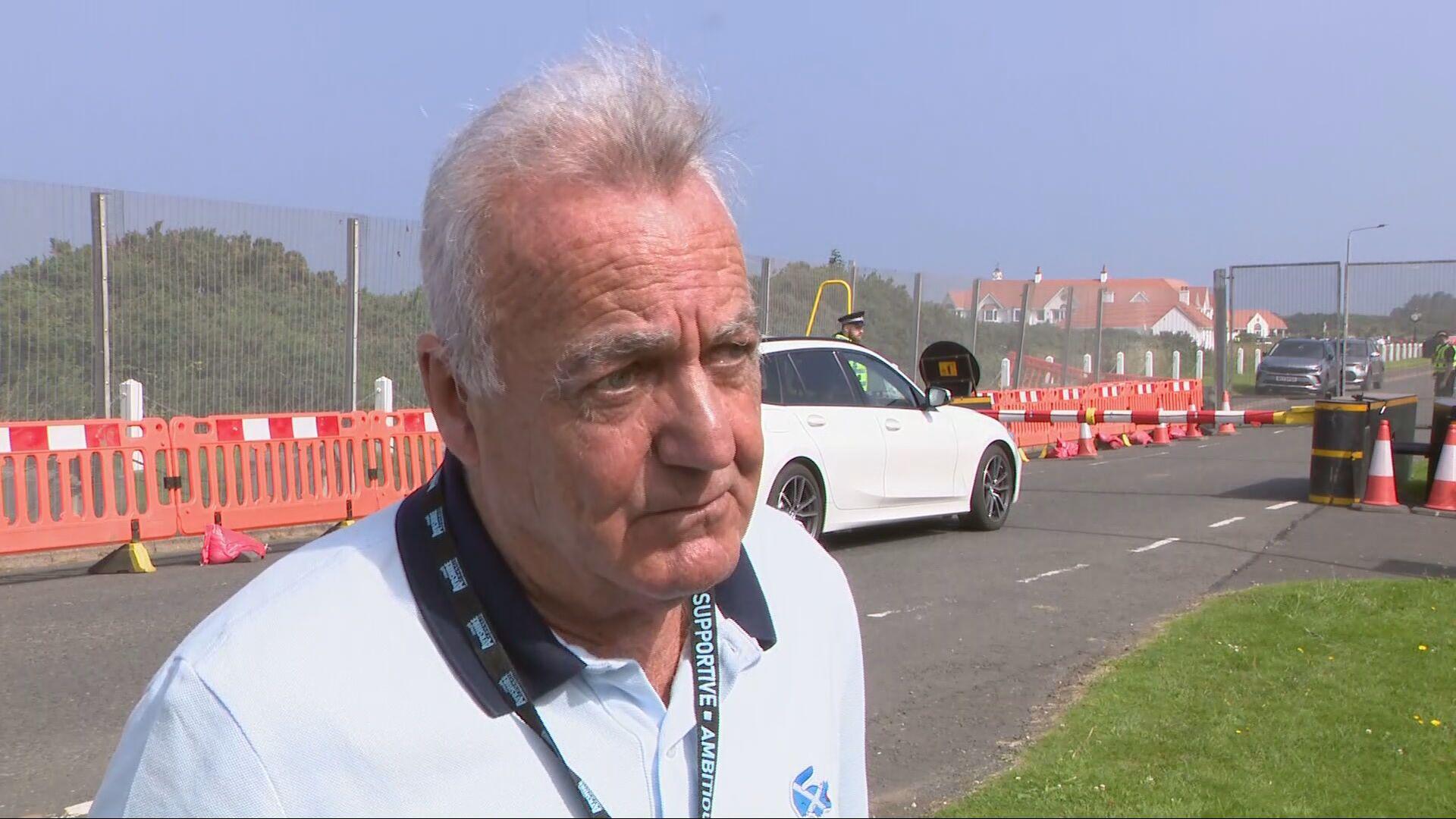 Gavin Scott looking slightly off camera. He has grey hair which is sticking up at the front. He is wearing a white polo shirt with a dark collar and a Saltire logo on the upper left chest. He is wearing a green lanyard around his neck with white writing on it.