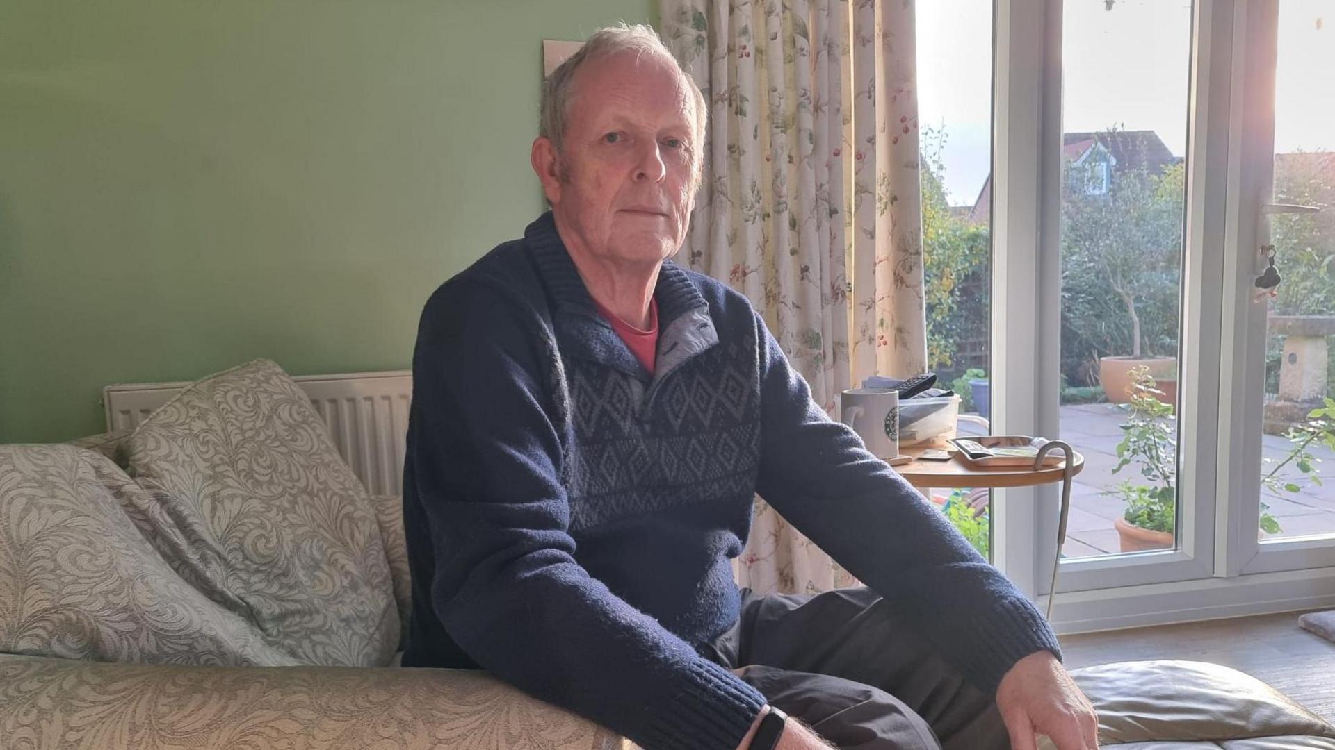 A man with white hair wearing a blue jumper sitting on a sofa. In the background is his house with windows out on a patio. 