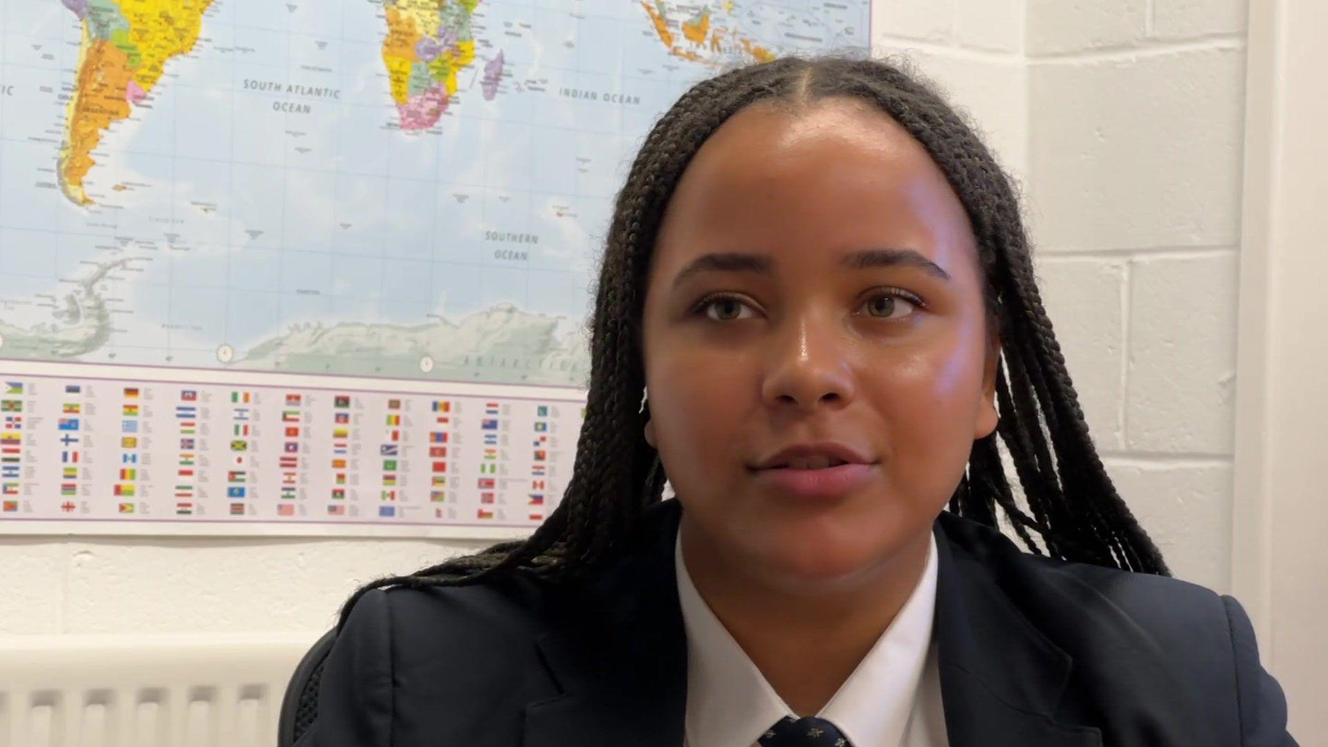 Inês sitting in a classroom in front of a map of the world, which hangs on the wall.