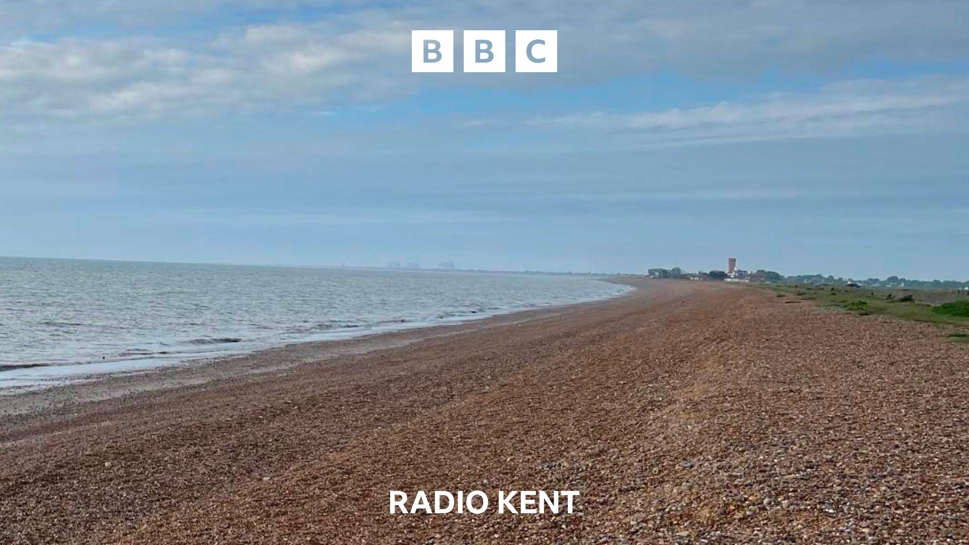 A shingle beach stretches off into the distance. The sky is blue with a small amount of cloud cover. The sea is calm.