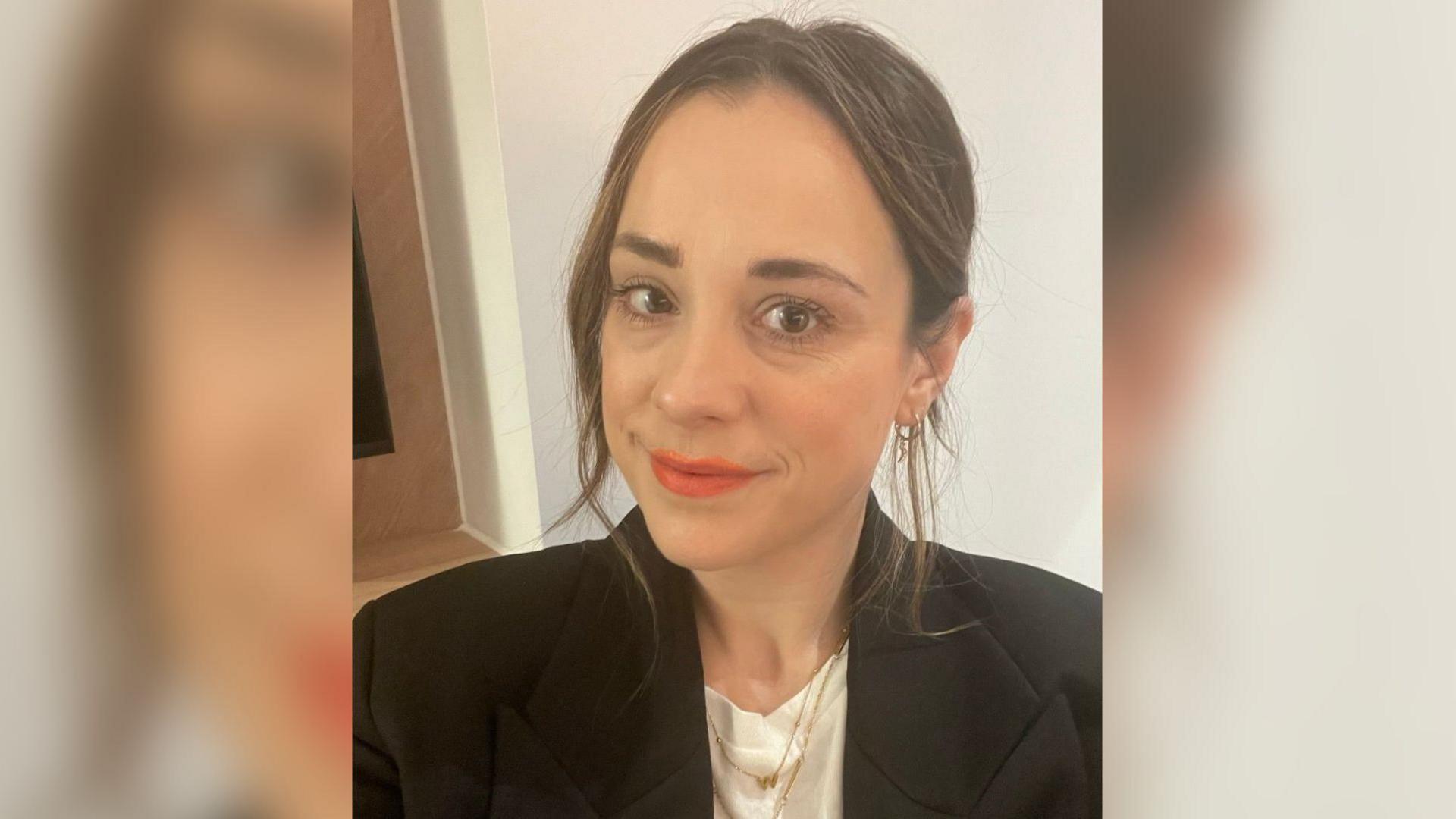 A head and shoulders image of a woman with long brown hair, tied back. She is wearing red lipstick. Her clothing comprises a black blazer with wipe lapels. Under this, she is wearing a white t-shirt.  