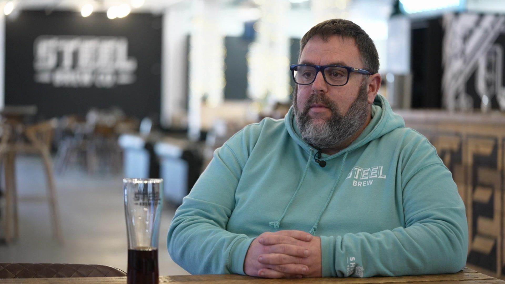 A man with a beard and glasses wearing a blue Steel Brew hoodie.