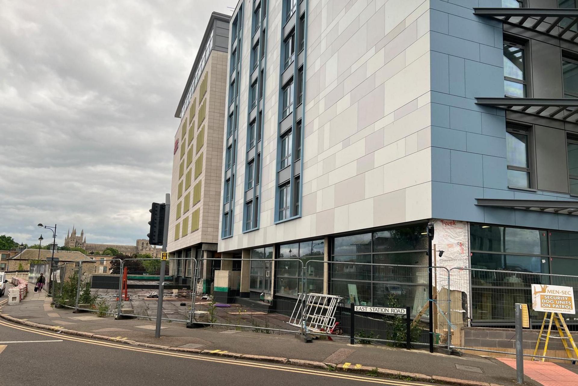 An unfinished hotel building. The grey exterior appears complete, but the ground floor windows show that it has not been fitted out. Metal fencing surrounds the site, with an EAST STATION ROAD street sign on the pavement and Peterborough Cathedral visible on the horizon.