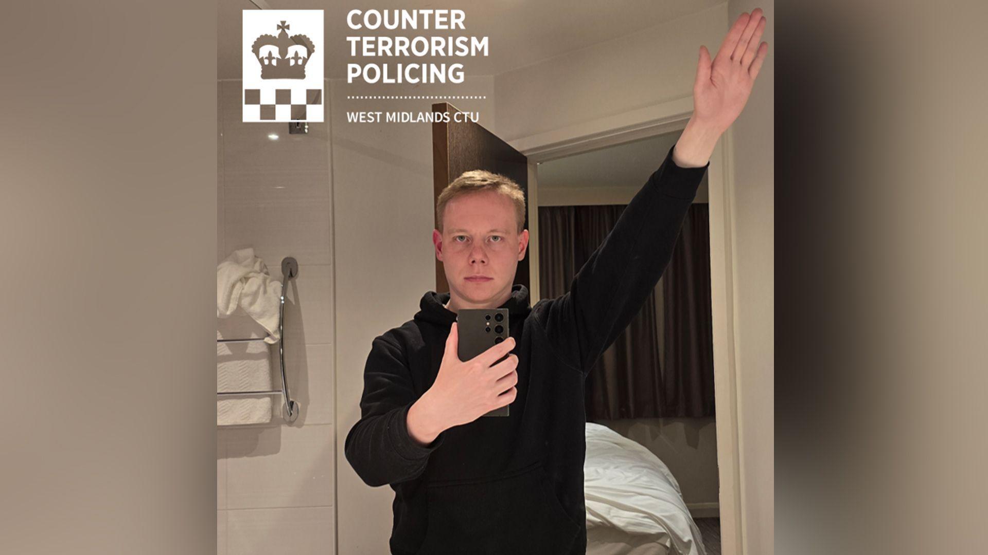 Callum Parslow, who has short blonde hair, wearing a black hoodie. He is taking a selfie in a mirror while making a Nazi salute with the other arm. Through the mirror image, you can see a bed and a towel rail in the background.