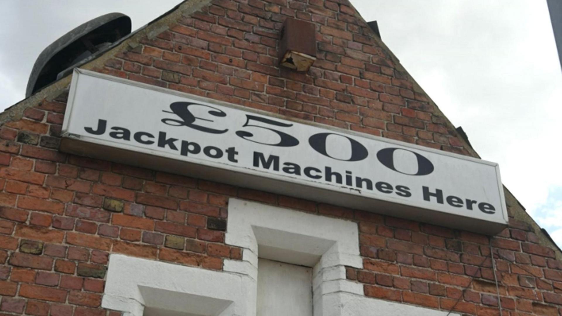 A white sign with black writing which states "£500 Jackpot Machines Here" on the side of a red brick building.