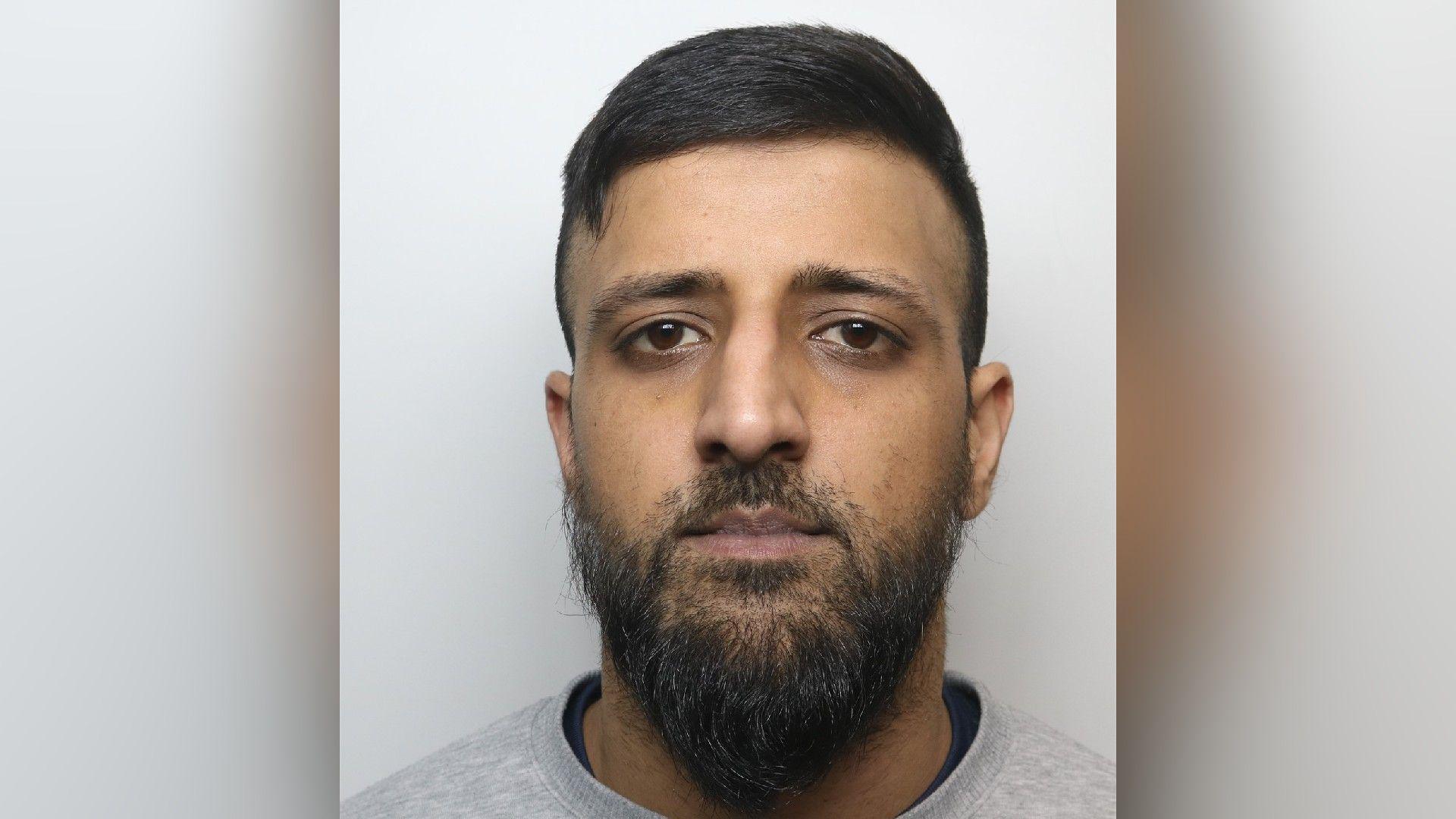 A mugshot of Shamrez Razaq who has black hair combed to one side and a medium length beard. He is wearing a grey jumper.