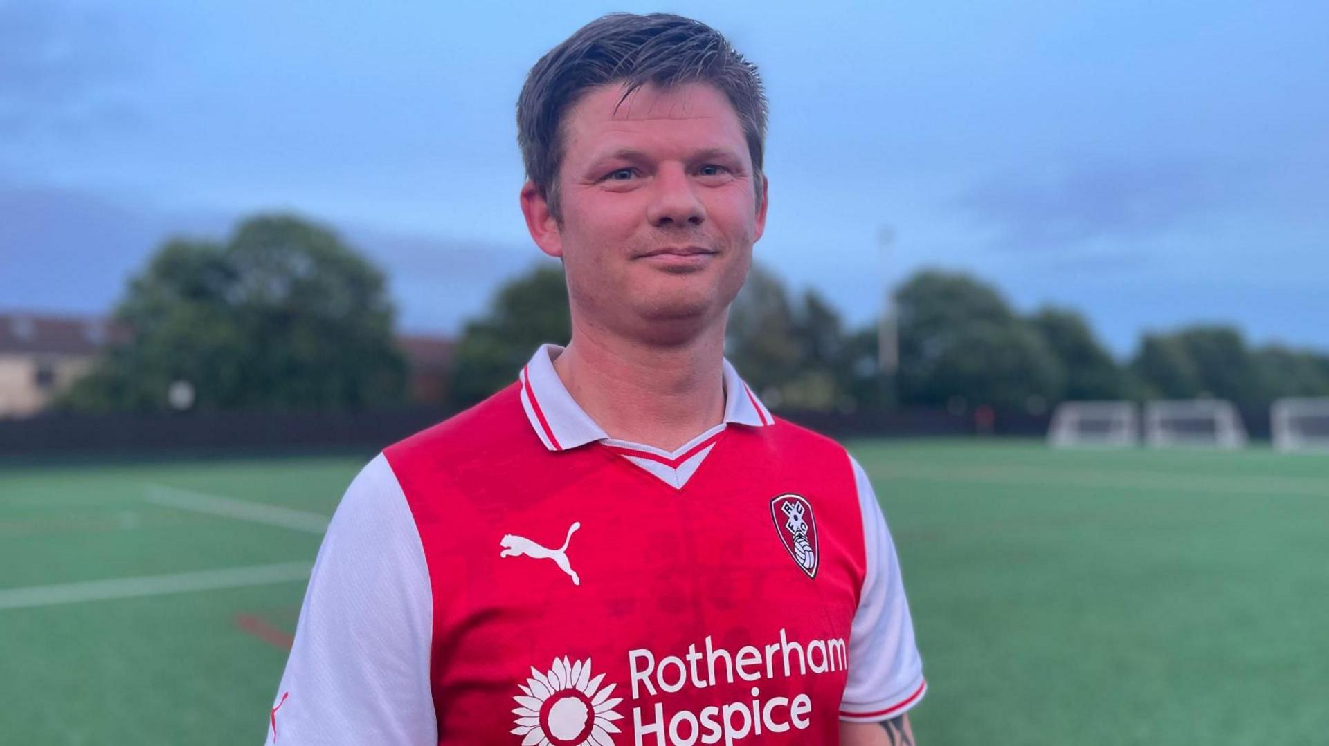A man with short dark hair stands on an empty football pitch wearing a red strip
