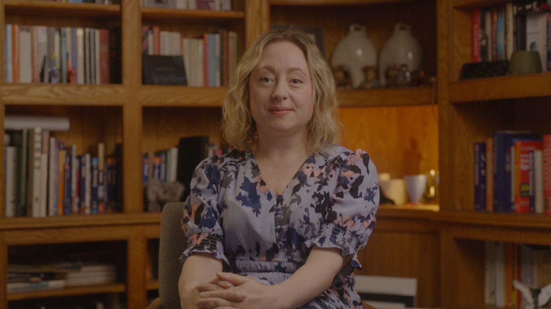 Photo of Catriona, seated, looking directly at the camera. She has shoulder-length blonde hair and is wearing a lavender-coloured pattern dress. She has her hands folded in her lap. Behind her are bookshelves containing books and other ornaments