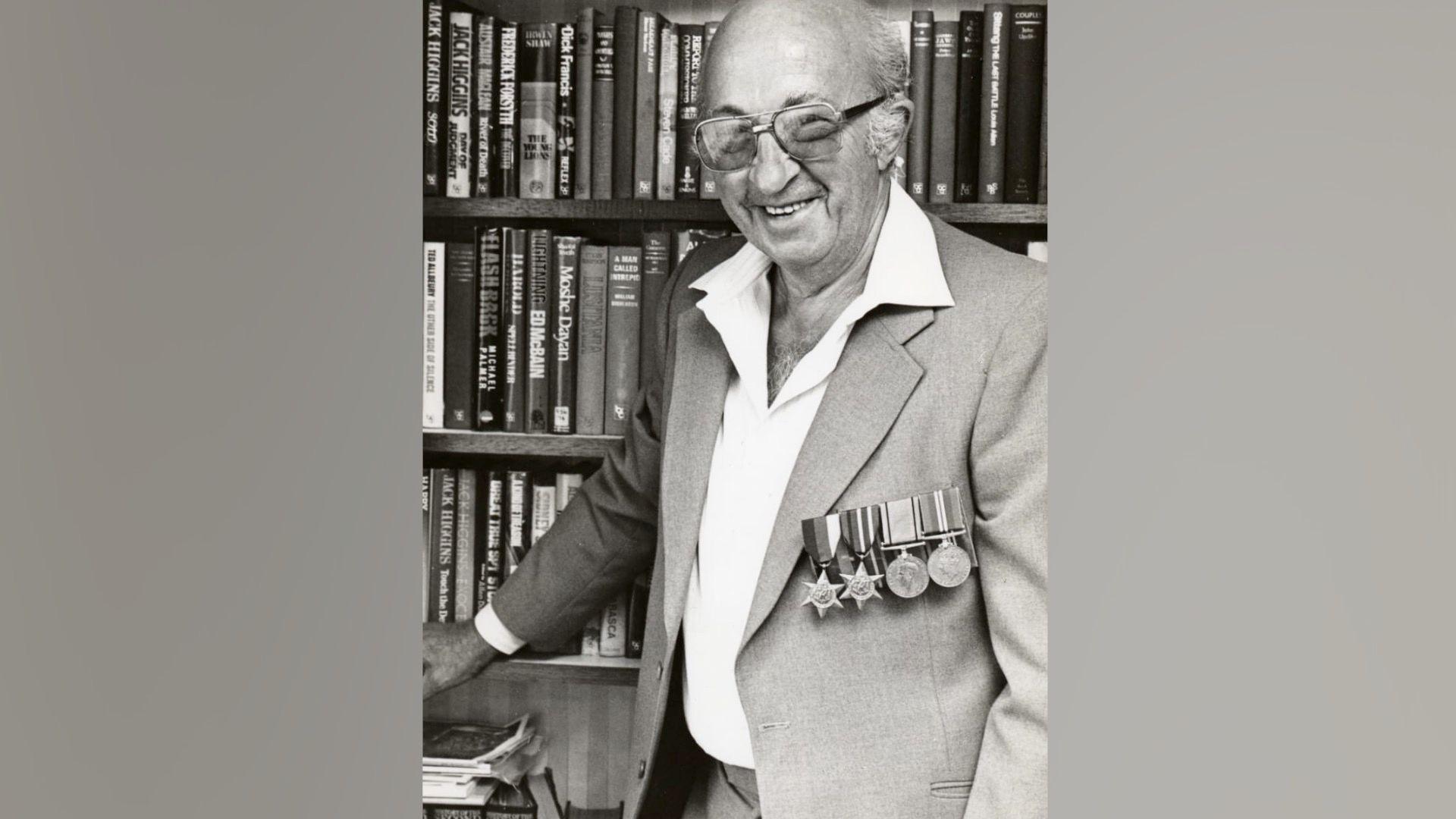 Alfonso Garizio is pictured in a black and white photograph. He is stood in front of a book case containing several books. He is wearing a grey suit jacket with white open-necked shirt underneath, and has four military medals attached to his left chest. He wears large-framed glasses and is smiling at the camera
