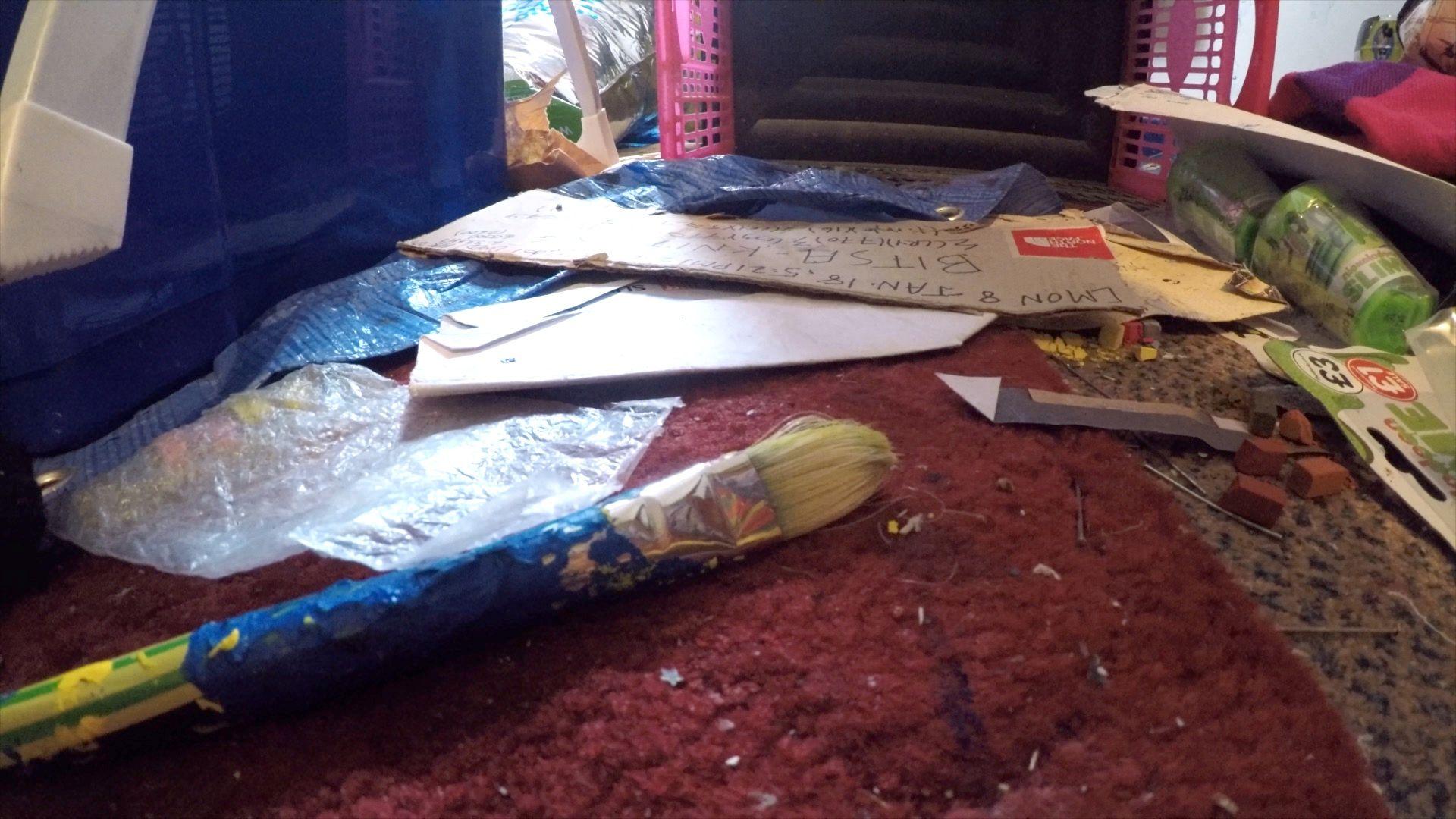 A close-up of the floor in one of Mr MacNee's rooms shows a red carpet with crumbs , nails and debris scattered across. A dirty paintbrush is in the foreground, with bits of paper and card behind it, two tubs of green slime, an upturned plastic basket and bits of plastic bubble wrap on the floor.