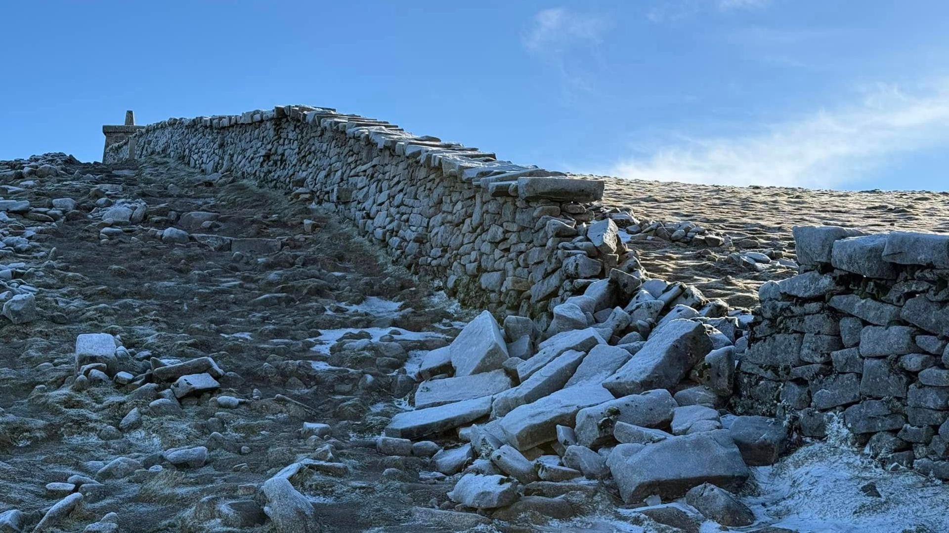 A granite stone wall rises up the side of a steep mountain. A section of the wall has collapsed and a large number of stones are lying in rubble.