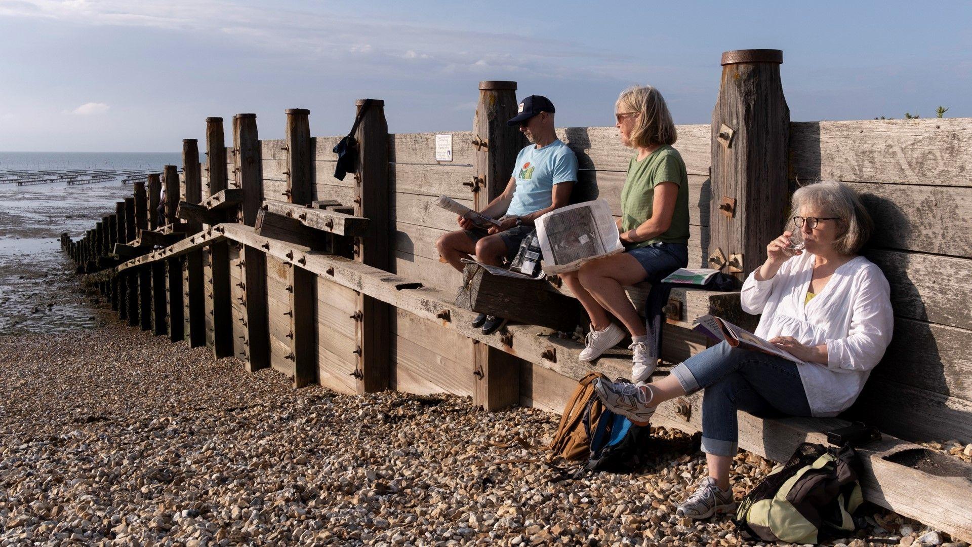 Three people sit above beach shingle on a wooden sea defence groyne, reading and drinking, during low-tide evening
