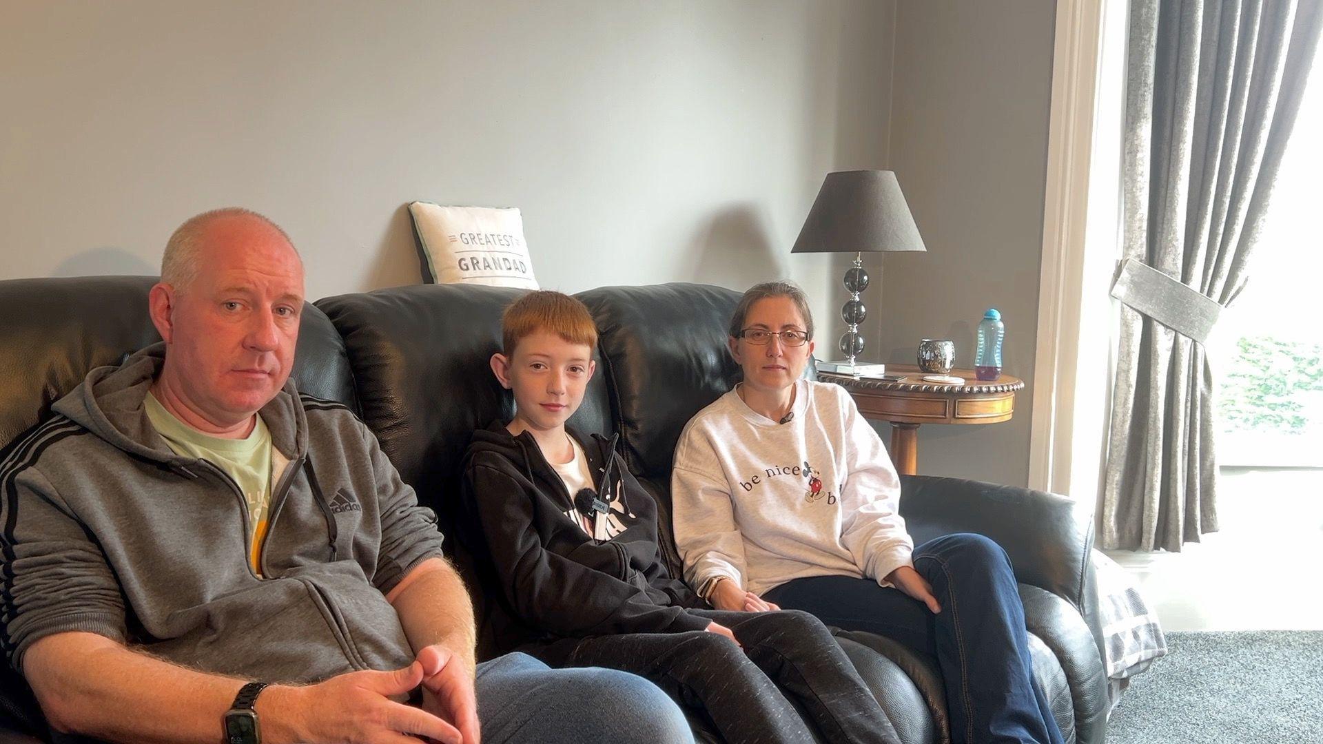 A man, woman and their 11-year-old son sit on a black leather sofa in a living room with grey walls, curtains, and carpet. A small brown coffee table is to the right of the sofa, with a lamp, candle and drink on it