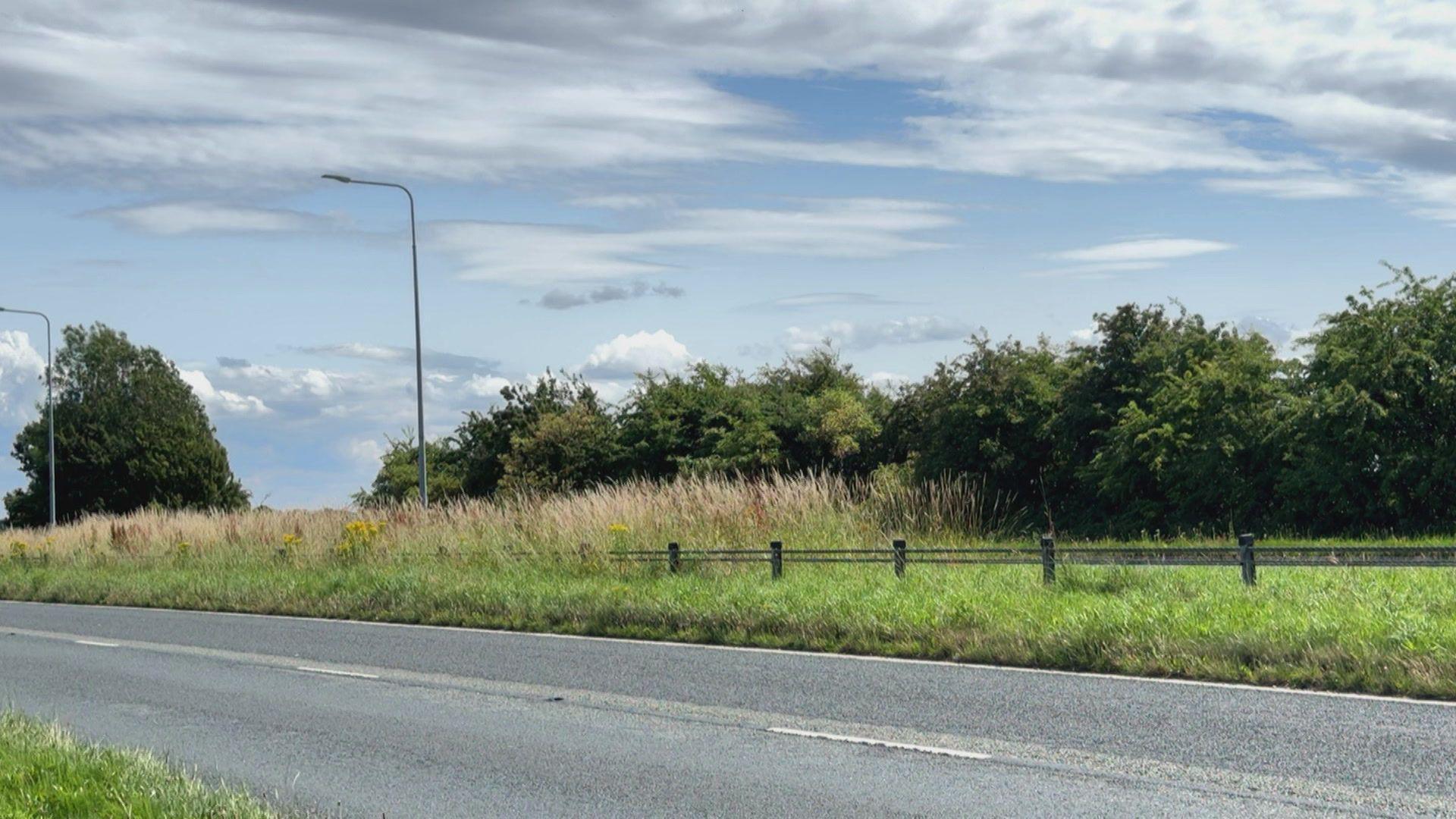 A picture of Rainford Road in Bickerstaff, with a grey two lane road in the foreground, a long grass verge in the background, with trees lining the road behind that. Two street lights can be seen on the left of the picture. The sky is blue with scattered cloud cover.
