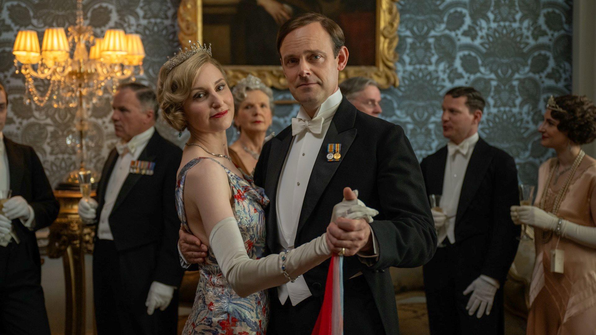 Lady Edith stands in her silk floral gown, looking at the camera, with the man who portrays her husband in the film. he is wearing a white shirt and black dinner jacket, they are dancing and looking at the camera.