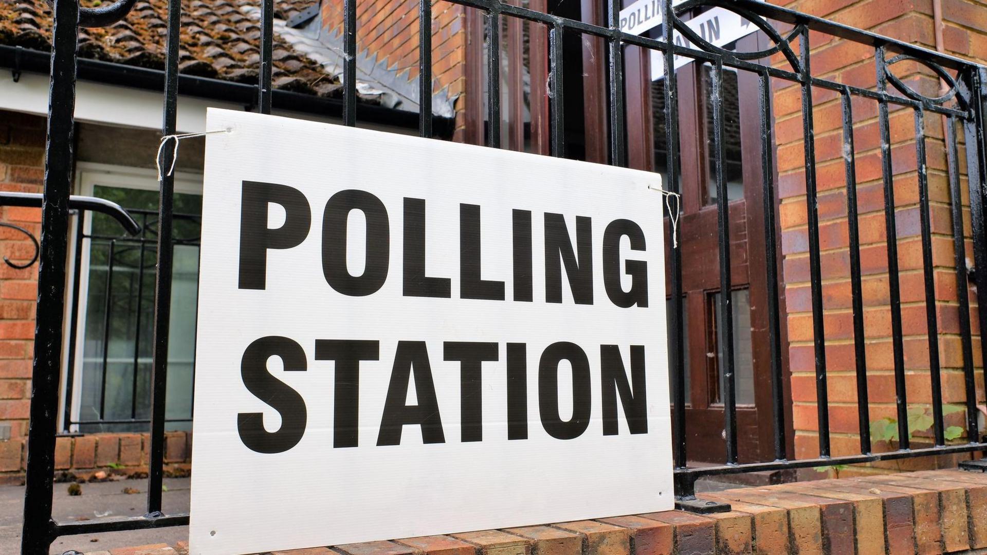 Red brick with a brown door behind railings and a big sign on the railings that reads 'POLLING STATION' in black capital letters against a white cardboard background. 